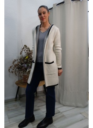 Modelo luciendo rebeca larga de punto para mujer en color blanco con ribetes negros en contraste, bolsillos frontales y capucha