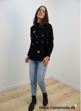 Modelo posando con jersey negro con detalles de pedrería brillantes y cuello redondo, combinado con jeans claros.