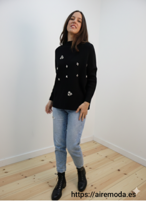 Modelo posando con jersey negro con detalles de pedrería brillantes y cuello redondo, combinado con jeans claros.