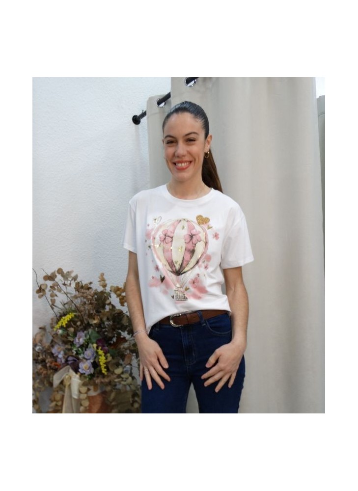 Modelo posando con camiseta blanca con ilustración de globo aerostático rosa y detalles de flores y corazones dorados