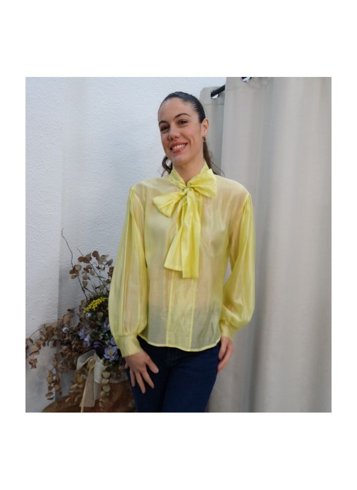 Modelo luciendo camisa de gasa semitransparente en color amarillo vibrante con lazo grande en el cuello y mangas abullonadas.